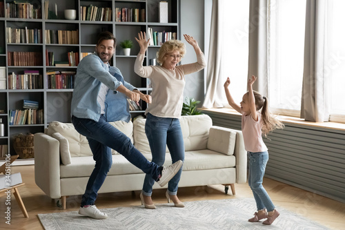 Little girl dancing in living room at home with father and elderly grand mother, funny multi-generational active family fooling around spending weekend time came to visit old granny having fun indoors