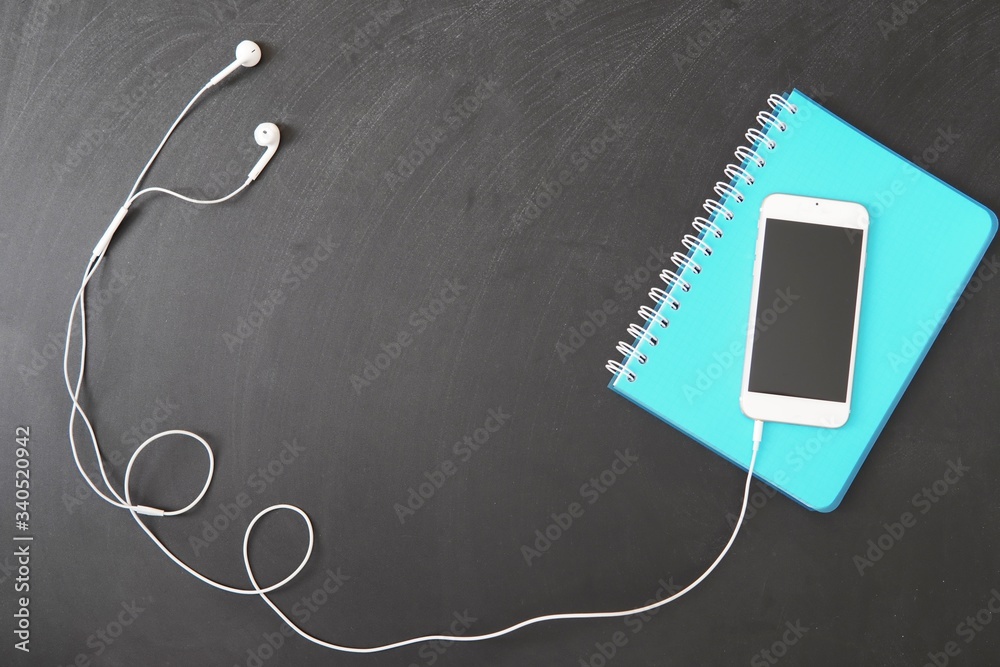 Remote learning background, smartphone with headphones and notebook on black chalkboard, online learning, distance education concept, space for text, top view. Remote learning background, smartphone with headphones and notebook on black chalkboard, online learning, distance education concept, space for text, top view.