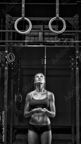 Slender young athlete getting ready for exercise on gymnastic rings. A girl in black sportswear is standing under the gymnastic rings in the gym, getting ready for working out. Black and white photo.
