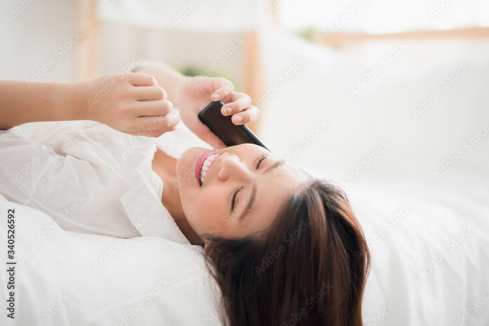 Young Asian woman lie supine which talking and using a smart phone on the white bed at home looking felling happy. the life at home concept, People work at home Young Asian woman lie supine which talking and using a smart phone on the white bed at home looking felling happy. the life at home concept, People work at home