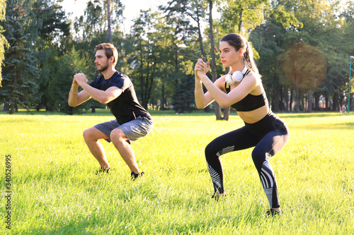 Young sporty man and woman doing workout and squatting together in green park during summer sunny day.