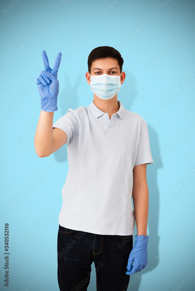 Teenage boy in medical mask and protective gloves showing victory gesture on blue background. Teen cured from covid-19. Positive concept of recovery, hope, confidence of victory over coronavirus. Teenage boy in medical mask and protective gloves showing victory gesture on blue background. Teen cured from covid-19. Positive concept of recovery, hope, confidence of victory over coronavirus.