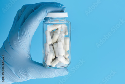 Hand in medicine glove holds a glass bottle with white capsule pills. Medicine pills in a bottle on blue background with copy space. Antiviral antibiotics in a glass container holding in hand.