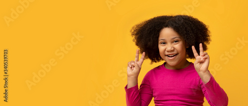 Joyful little afro girl gesturing peace sign and smiling at camera