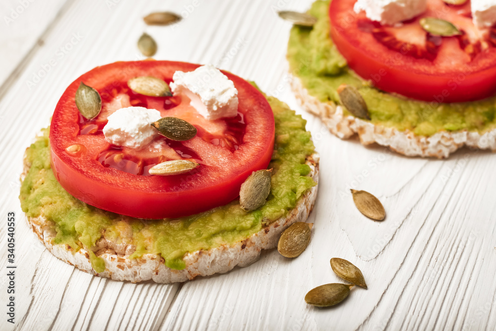 Rice cake with avocado and tomatoes and feta cheese with pumpkin seeds for breakfast on a white wooden background