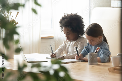 Concentrated small multiracial sisters sit at the desk having online class on laptop, little multiethnic girls siblings study together engaged in web lesson on computer at home, homeschooling concept