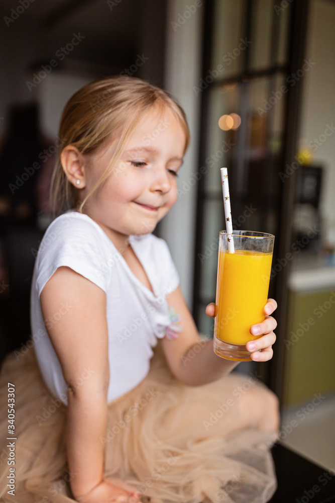 Cute little caucasian girl with blonde hair sitting on the kitchen table and drinking fresh mango juice. Healthy lifestyle concept. Kid 6 years old at home Cute little caucasian girl with blonde hair sitting on the kitchen table and drinking fresh mango juice. Healthy lifestyle concept. Kid 6 years old at home