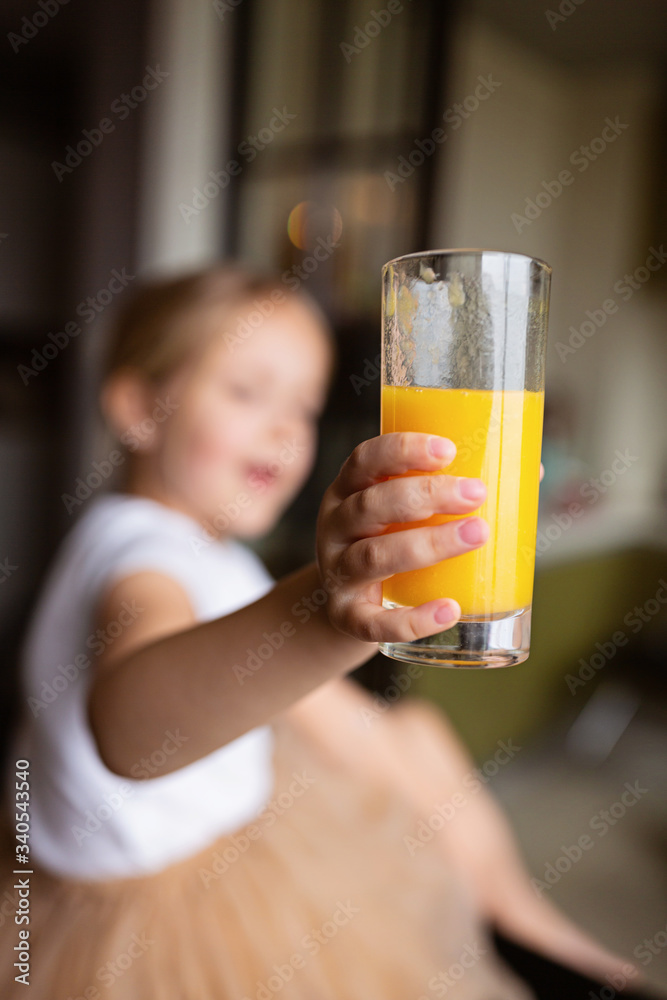 Cute little caucasian girl with blonde hair sitting on the kitchen table and drinking fresh mango juice. Healthy lifestyle concept. Kid 6 years old at home Cute little caucasian girl with blonde hair sitting on the kitchen table and drinking fresh mango juice. Healthy lifestyle concept. Kid 6 years old at home