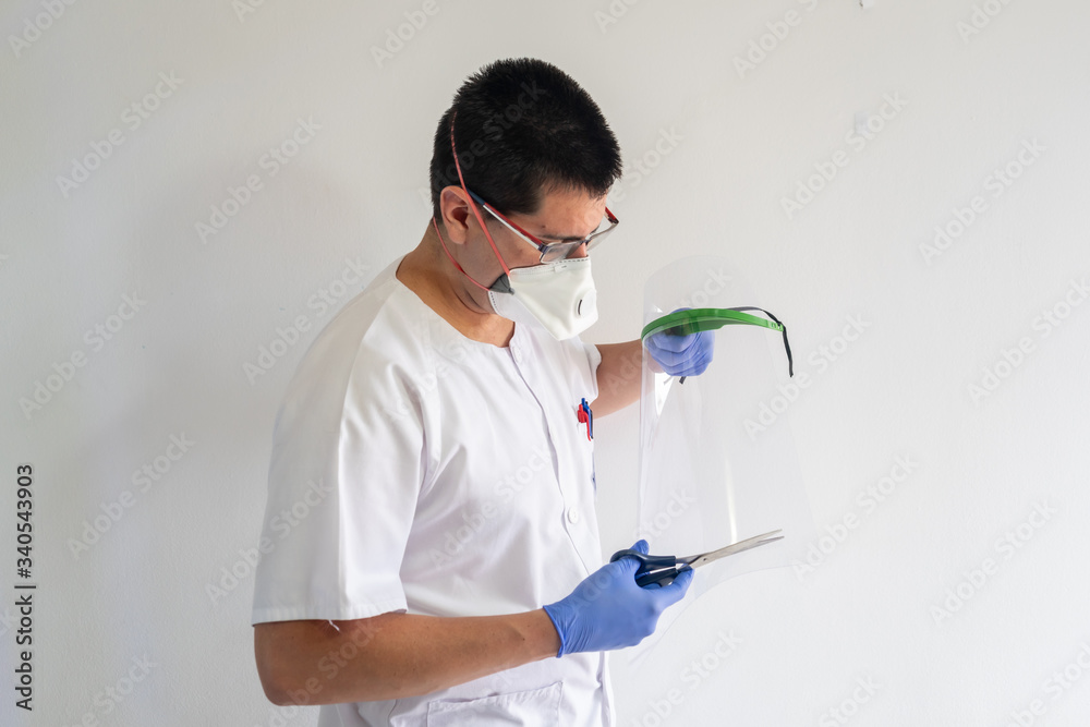 Naklejka premium Doctor / nurse cutting a face shield with scissors to protect against the coronavirus. He is a young man. He wears gloves, masks, and glasses. He wears a white uniform.