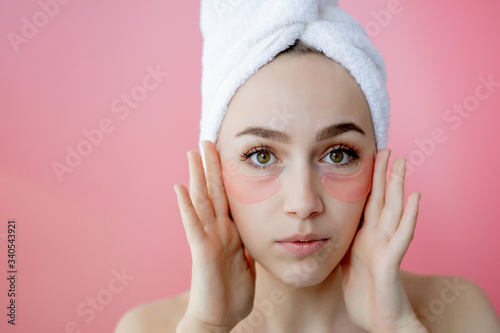 Studio shot of satisfied caucasian freckled woman wearing white towel on head, with collagen patches under eyes, standing naked against pink background. Skin care, cosmetic product concept