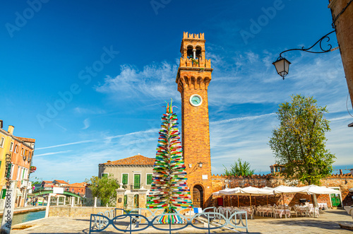 Murano clock tower Torre dell'Orologio of San Stefano church and Colorful christmas tree made of Murano Glass on Campo Santo Stefano square in Murano islands, Veneto Region, Northern Italy photo