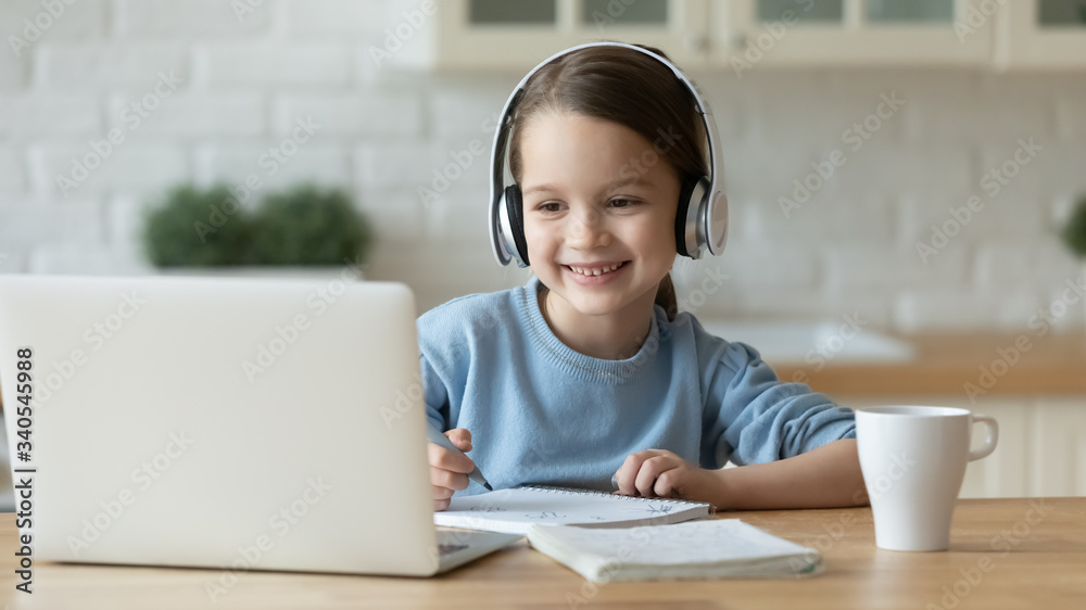 Smiling little Caucasian girl in headphones handwrite study online using laptop at home, cute happy small child in earphones take Internet web lesson or class on computer, homeschooling concept - obrazy, fototapety, plakaty 