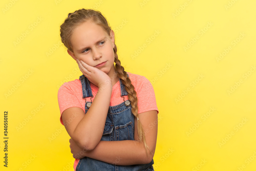 Portrait of pensive thoughtful little girl with braid in denim overalls leaning on hand, pondering answer with serious expression, thinking over idea. indoor studio shot isolated on yellow background Portrait of pensive thoughtful little girl with braid in denim overalls leaning on hand, pondering answer with serious expression, thinking over idea. indoor studio shot isolated on yellow background