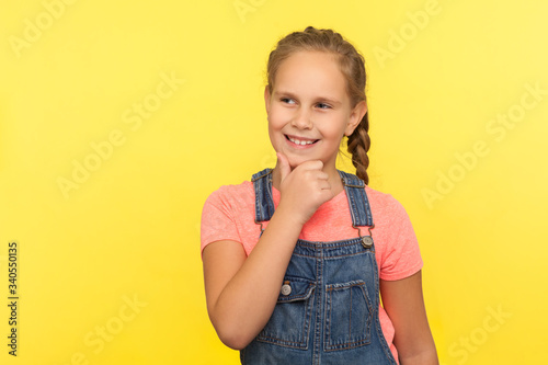 Portrait of positive thoughtful little girl in denim overalls holding chin and dreaming with cunning expression, fantasizing, having interesting idea. studio shot isolated on yellow background