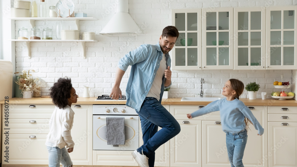 Obraz premium Overjoyed young Caucasian father and little multiracial daughters dancing together in home kitchen, happy dad and small multiethnic girls children have fun playing on weekend or quarantine