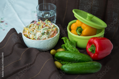Decorated with lettuce, side view, selective focus . Orange pepper in a green bowl . Water in a glass. Brown tablecloth. Salad with ham, bell pepper, mushrooms, pickles, white sauce, on a white plate.
