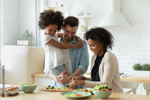 Happy young diverse parents have fun teach little biracial daughter cooking  overjoyed multiracial family with small girl child preparing food making healthy salad for breakfast in kitchen together