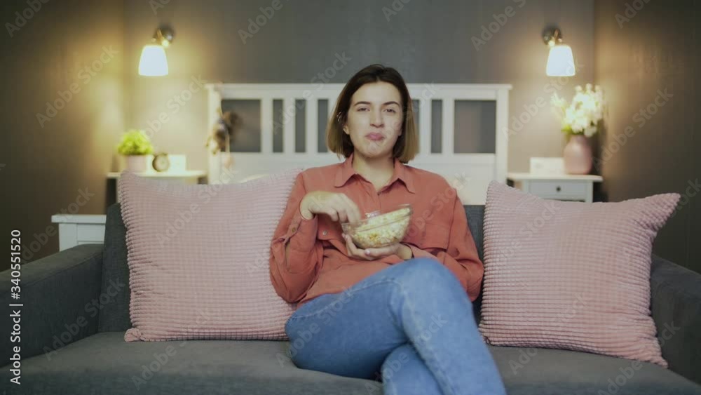 Happy young woman sitting on the sofa, eating popcorn, watching TV and laughing