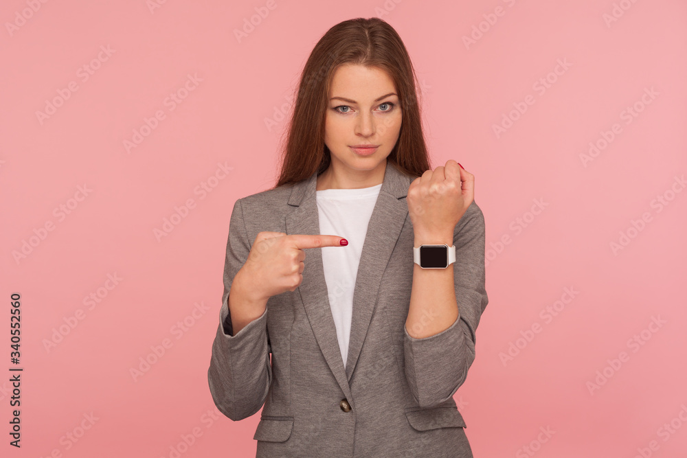 It's late, hurry up! Portrait of impatient worried young busy lady in business suit pointing to watch on her wrist and looking displeased, anxious about deadline, delay. indoor studio shot, isolated
