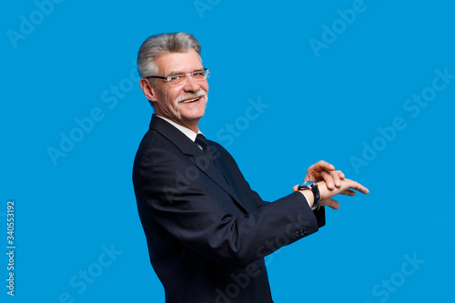 Photo of a handsome smiling older businessman dressed inblack suit  standing isolated over blue background, looking at hand watch and smiling photo