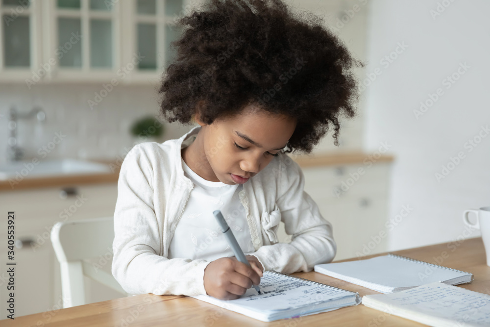 Small African American girl sit at desk in kitchen handwrite in notebook having distant class, smart little biracial kid prepare homework study alone on quarantine at home, homeschooling concept Small African American girl sit at desk in kitchen handwrite in notebook having distant class, smart little biracial kid prepare homework study alone on quarantine at home, homeschooling concept