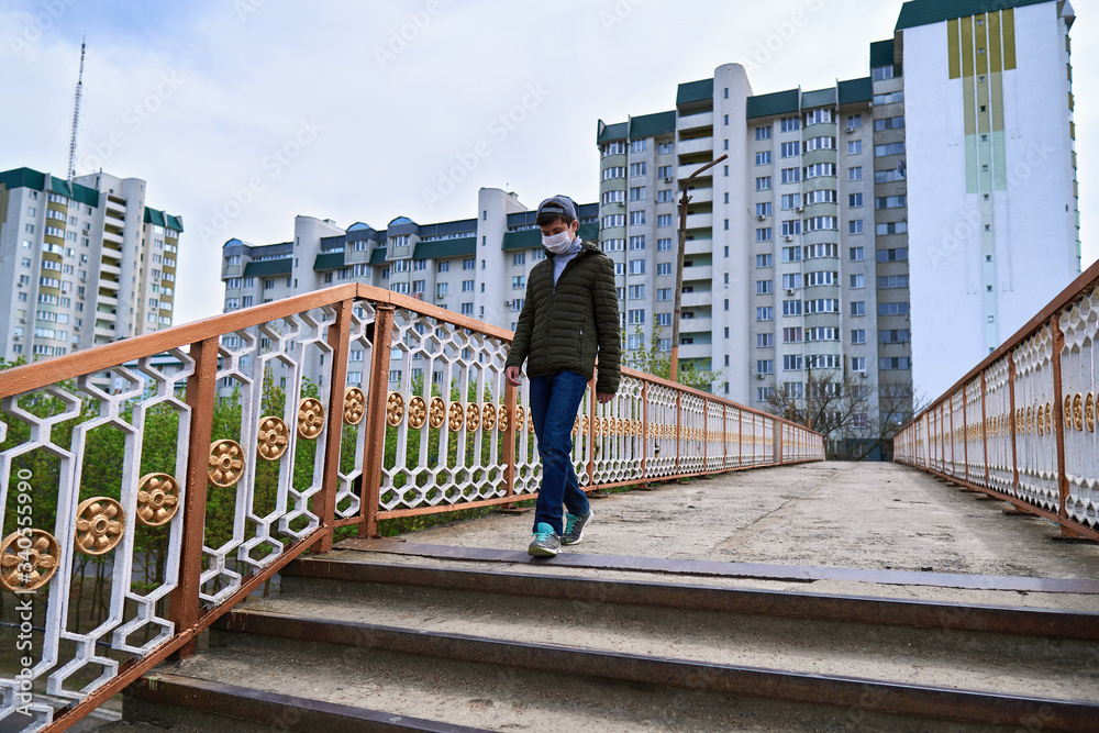 teen boy walks down the street during the day, a pedestrian walkway and high-rise buildings with apartments, a residential area, a medical mask on his face protects against viruses and dust teen boy walks down the street during the day, a pedestrian walkway and high-rise buildings with apartments, a residential area, a medical mask on his face protects against viruses and dust
