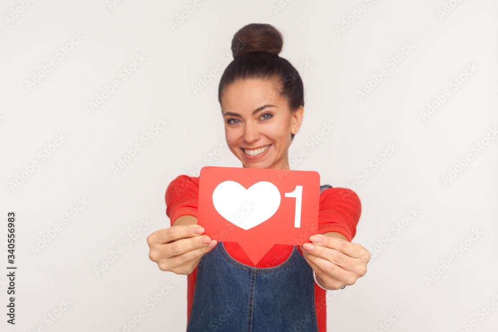 Like and follow interesting content! Portrait of charming positive trendy girl with hair bun in denim overalls showing social media heart icon to camera and smiling. indoor studio shot, isolated Like and follow interesting content! Portrait of charming positive trendy girl with hair bun in denim overalls showing social media heart icon to camera and smiling. indoor studio shot, isolated