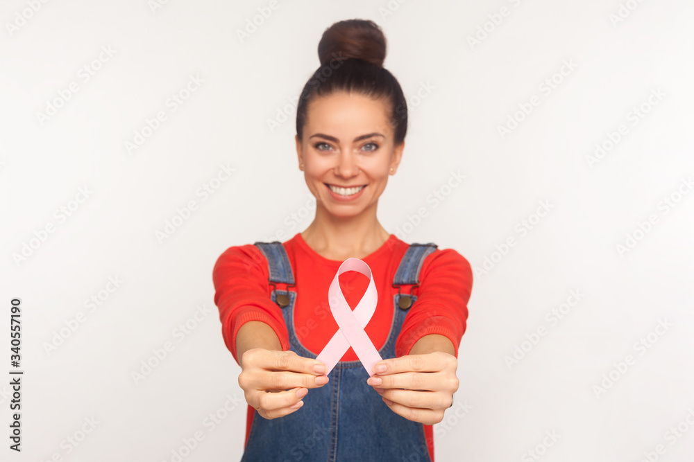Breast cancer awareness. Optimistic happy girl with hair bun in casual style denim overalls smiling and showing pink ribbon, symbol of female health. indoor studio shot isolated on white background Breast cancer awareness. Optimistic happy girl with hair bun in casual style denim overalls smiling and showing pink ribbon, symbol of female health. indoor studio shot isolated on white background