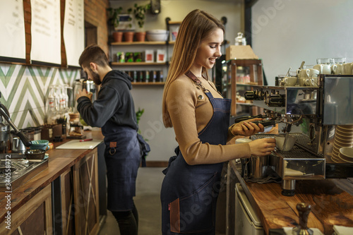 Professional female barista holding metal jug warming milk using the coffee machine. Positive smiling woman preparing coffee at machine © Тарас Нагирняк