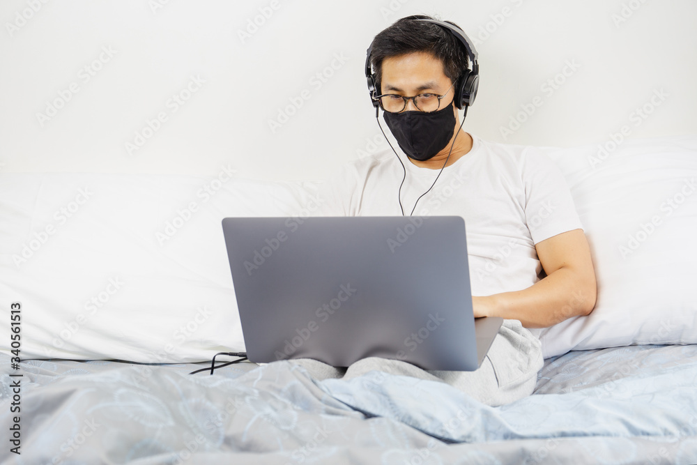 Man with protective mask and headphone working on laptop in bed at home covid-19 outbreak. Man with protective mask and headphone working on laptop in bed at home covid-19 outbreak.