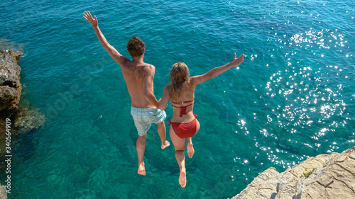 Joyful tourist couple decides to jump off a rocky cliff and dive into sea.