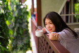 young happy and beautiful Asian Korean woman having morning coffee or tea at hotel terrace or home balcony leaning on the balustrade relaxed and cheerful