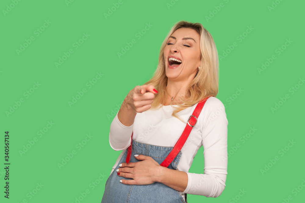 Ha ha ha, look at you! Portrait of adult woman bursting into laughing while watching someone really funny and ridiculous, pointing finger to camera. indoor studio shot isolated on green background