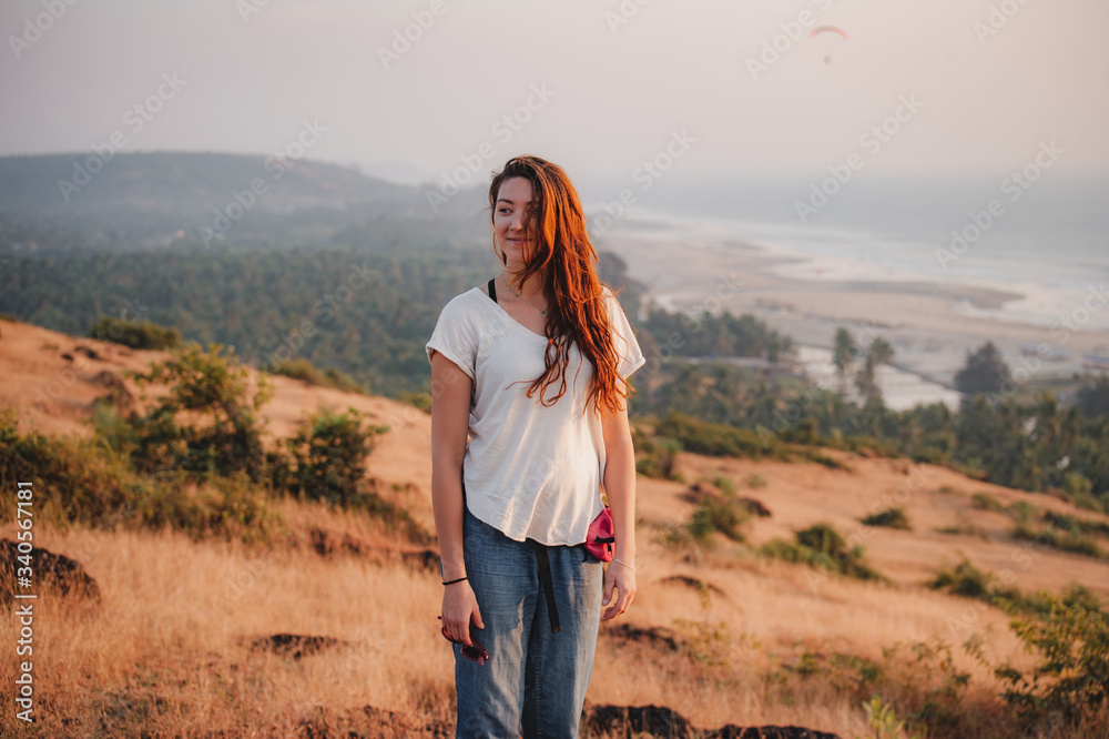 A girl with red hair, in a white T-shirt and blue jeans, with sunglasses in her hand, stands with a smile on top of a hill against the background of the ocean