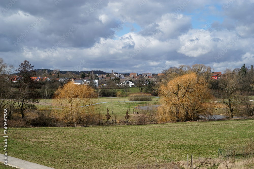 Landschaft mit Bäumen im Vordergrund und Wohnhäusern im Hintergrund bei bewölktem Himmel im Frühling. Bayreuth. Deutschland. Erholungsgebiet Mainaue. 