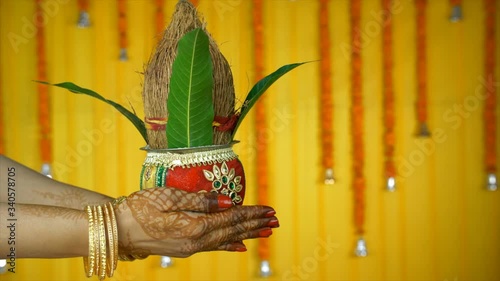 Side view shot of henna mehndi/tattoo on woman hands holding a wedding Kalash. Indian Pooja. Closeup of Indian female showing a Puja Kalash with coconut and mango leaves against a beautiful floral ... photo