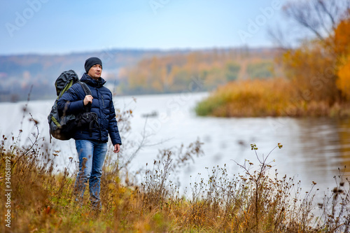 A young man with rucksack standing next to the river. Good warm autumn weather for hiking. © Vadim