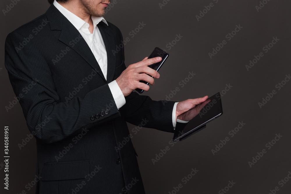 Businessman in black suit holding tablet pc in left hand and smartphone in right hand closeup side view. Showing screen to screen mobile to a tablet. Businessman in black suit holding tablet pc in left hand and smartphone in right hand closeup side view. Showing screen to screen mobile to a tablet.