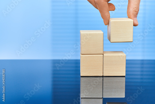 Four wooden cubes on a blue background in two rows with a reflection on the surface. A man's hand raises the upper cube on the left. Free space for letters, numbers, symbols, or labels.