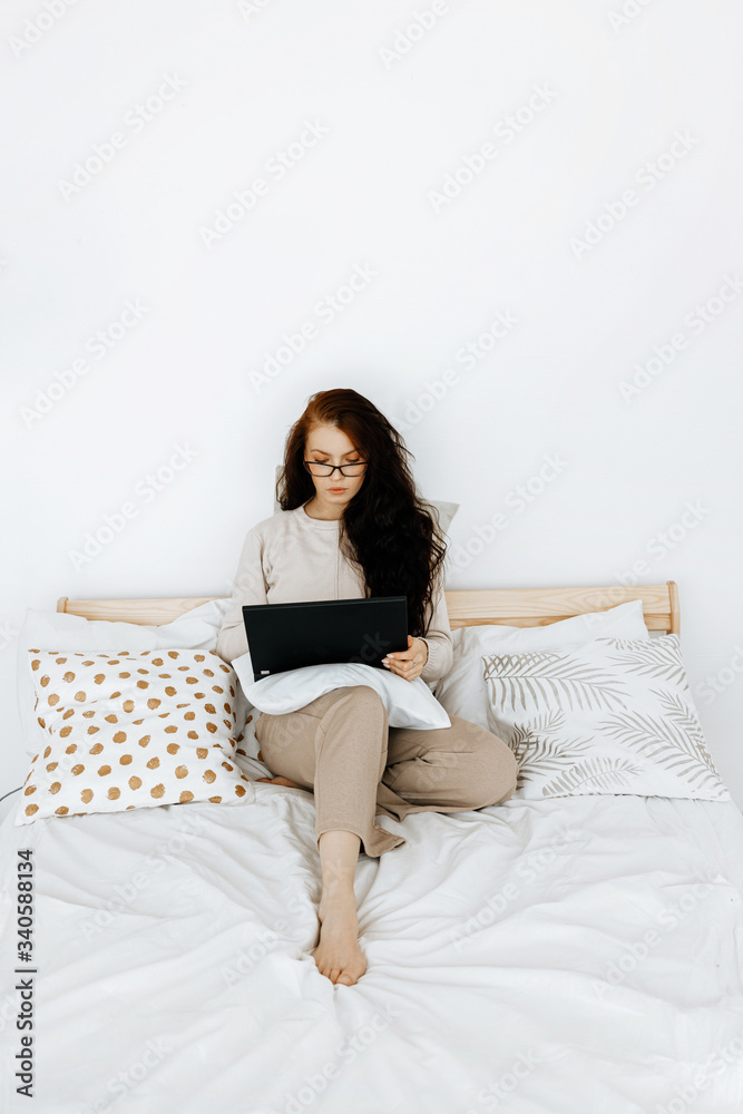 Distance learning or working from home concept. A young beautiful woman in beige home clothes is studying or working from home on a laptop in her bedroom via the Internet. Distance learning or working from home concept. A young beautiful woman in beige home clothes is studying or working from home on a laptop in her bedroom via the Internet.
