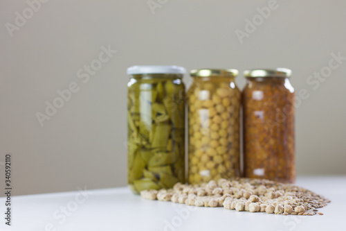 Photo of non-perishable food purchased due to Covid-19, chickpeas, lentils, and green beans in jars and spilled on white wooden board. 