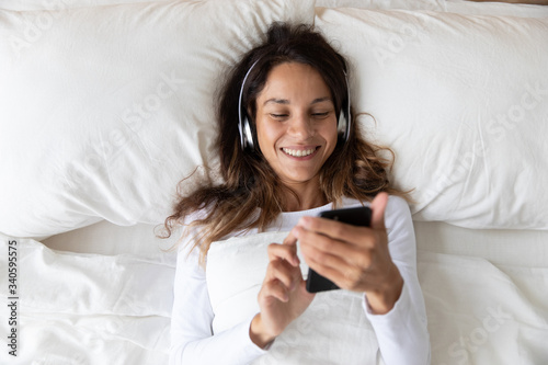 Top view close up smiling woman wearing headphones enjoying music, holding phone, resting on soft pillow in bedroom in morning, happy beautiful girl listening to favorite popular song, lying in bed