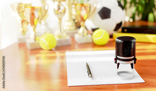 Office workplace with a blank sheet, pen, stamp, soccer and tennis balls, trophies on a blurred background. Sports business concept. Management, negotiations, contracts. Copy space. Selective focus. photo