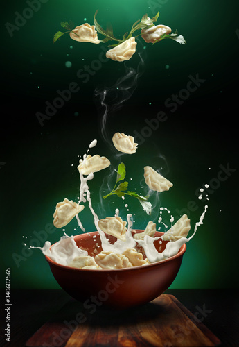 Hot pierogi flying out of the clay bowl with cream and parsley. Some vareniki stay inside the plate.  Green background.