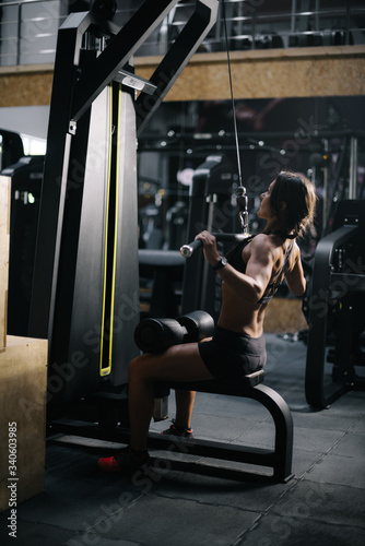 Strong unrecognizable sporty woman using training simulator for pumping back muscles in gym, back of view. Concept of professional training in a modern dark gym.