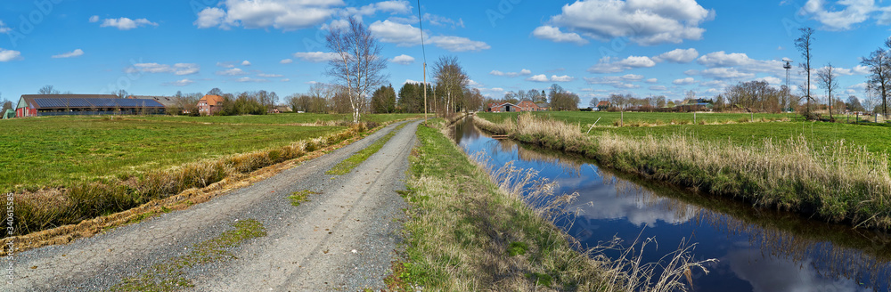 panorama view of typical marsh landscape in the district Wesermarsch (Germany) with green meadows, an unfixed rural lane, a canal and farm houses under vivid blue sky with white clouds in sunny spring panorama view of typical marsh landscape in the district Wesermarsch (Germany) with green meadows, an unfixed rural lane, a canal and farm houses under vivid blue sky with white clouds in sunny spring
