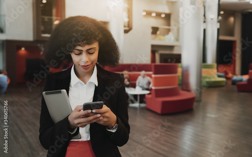 Portrait of an African-American woman entrepreneur standing indoors in office open-space area and typing a message on a smartphone with a digital tablet in another hand; a copy space place on the left