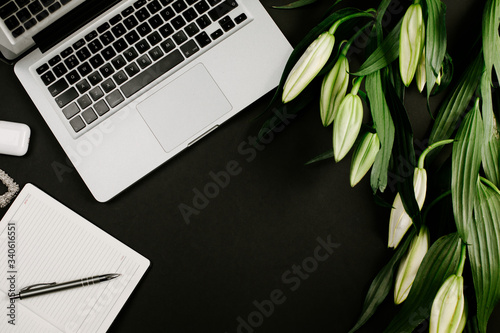 Modern workspace with laptop keyboard, notebook, pen and green flowers on black desk. Top view, flat lay