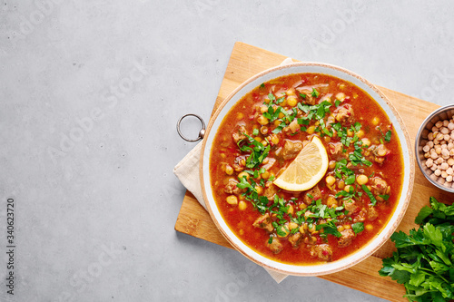 Moroccan Harira Soup in white plate on woden board on grey concrete table top. Harira is Moroccan Cuisine dish with lamb or beef meat, chickpeas, lentils, tomatoes, ciliantro. Ramadan Iftar Food.