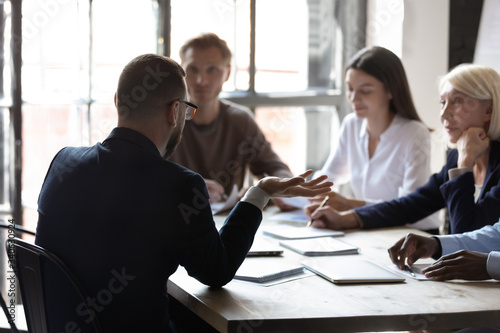 Back view of male employee talk with diverse multicultural colleagues at office briefing, businessman speak brainstorm with multiethnic coworkers or business partners at meeting, cooperation concept